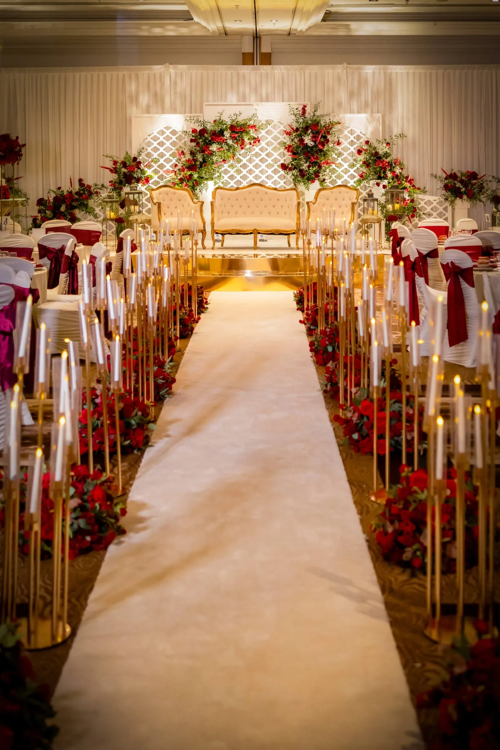 A red and gold wedding stage by Diamond Events: tufted gold settees beneath red rose arrangements and a white lattice